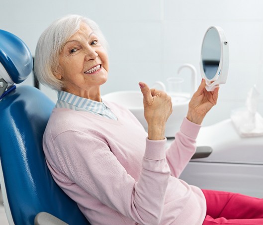 Senior woman sitting in dental chair giving thumbs up while holding a mirror